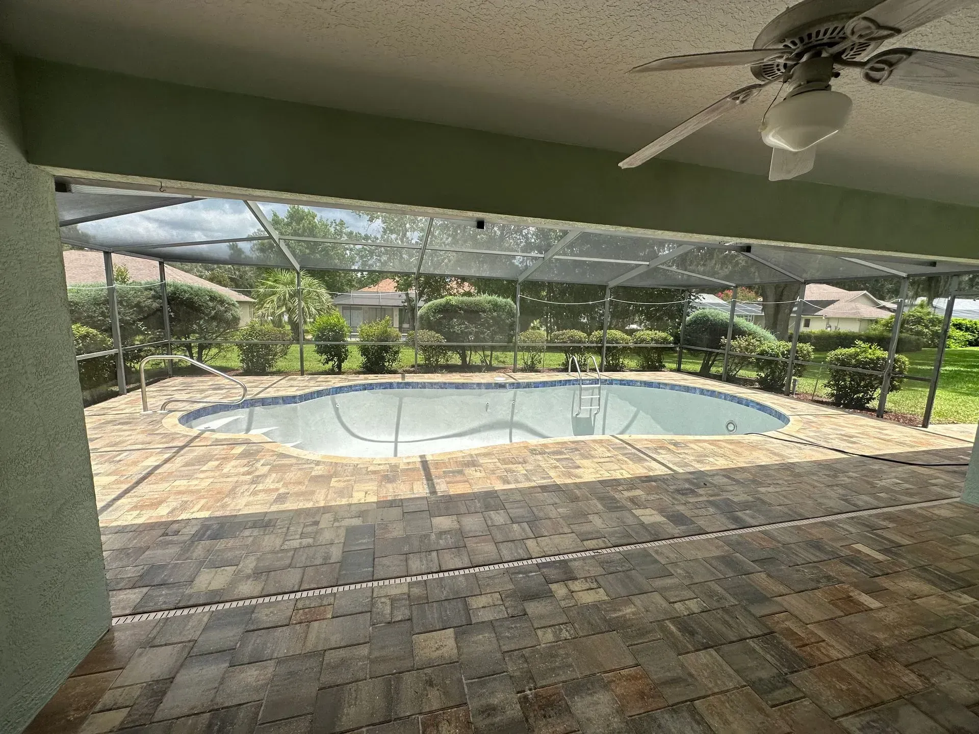 Patio with brick pavers, screened-in pool, and lush green landscaping. A ceiling fan is visible above.