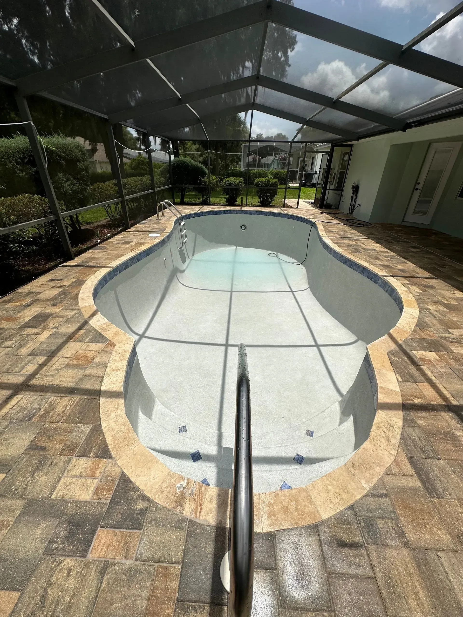 Empty swimming pool surrounded by brick pavers, under a screened enclosure, with a handrail.