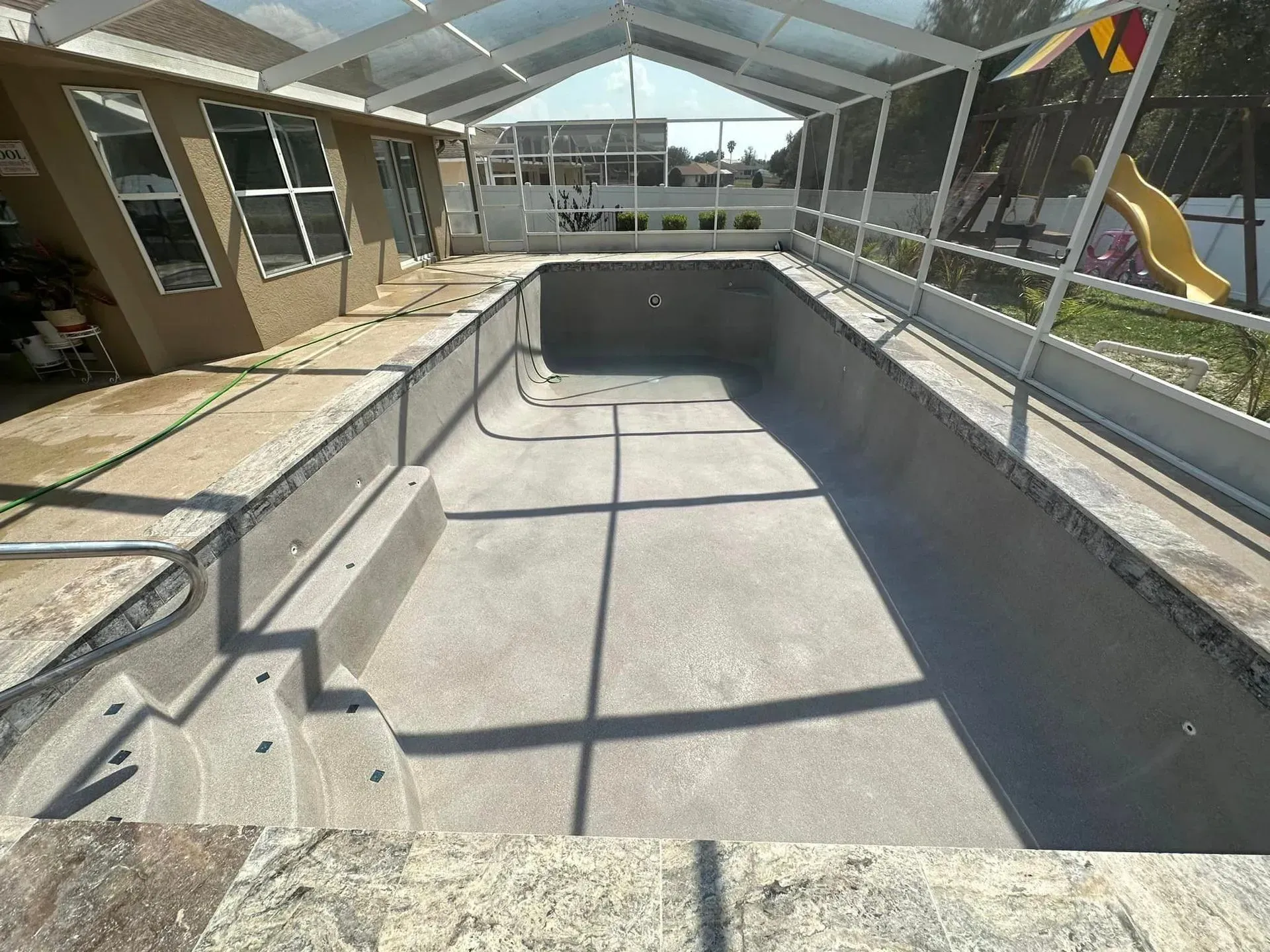 Empty swimming pool under a screen enclosure; concrete structure, steps, and surrounding patio.