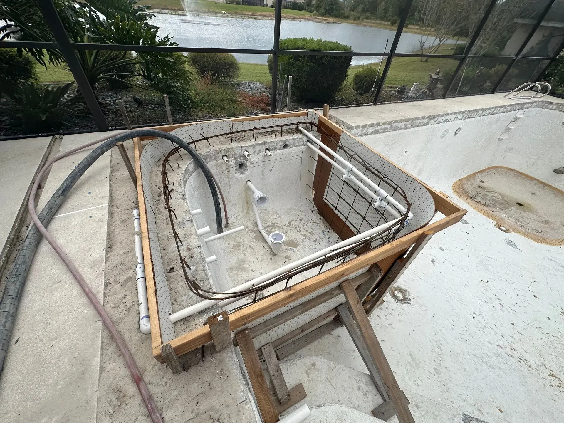 Pool undergoing renovation, showing the structure with exposed pipes, framing, and concrete. Outdoors with a pond view.
