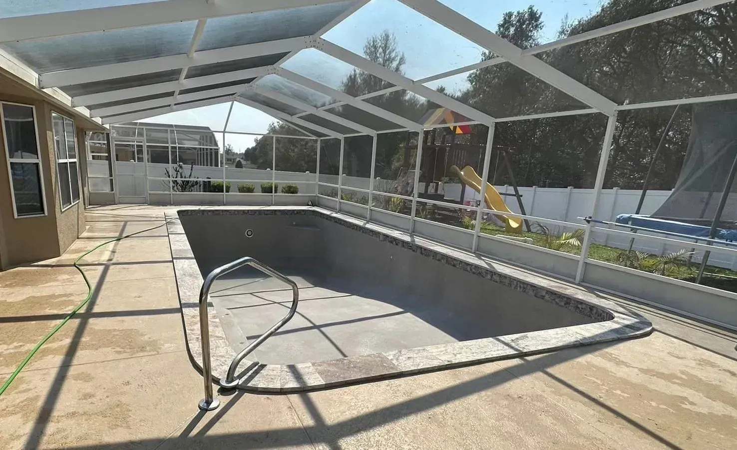 Empty rectangular pool under a white-framed screen enclosure, concrete deck, backyard with trees and a slide.