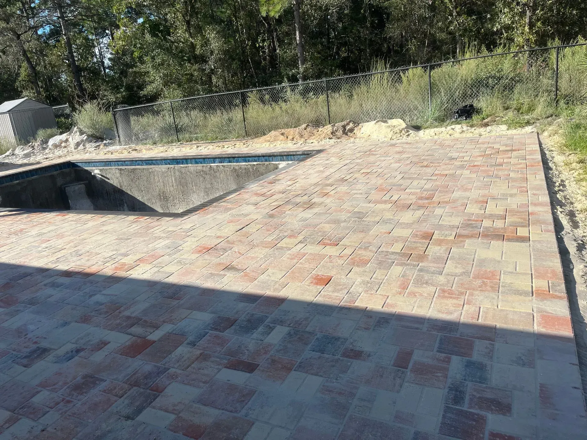 Brick patio next to a pool under construction, trees in the background, sunny day.
