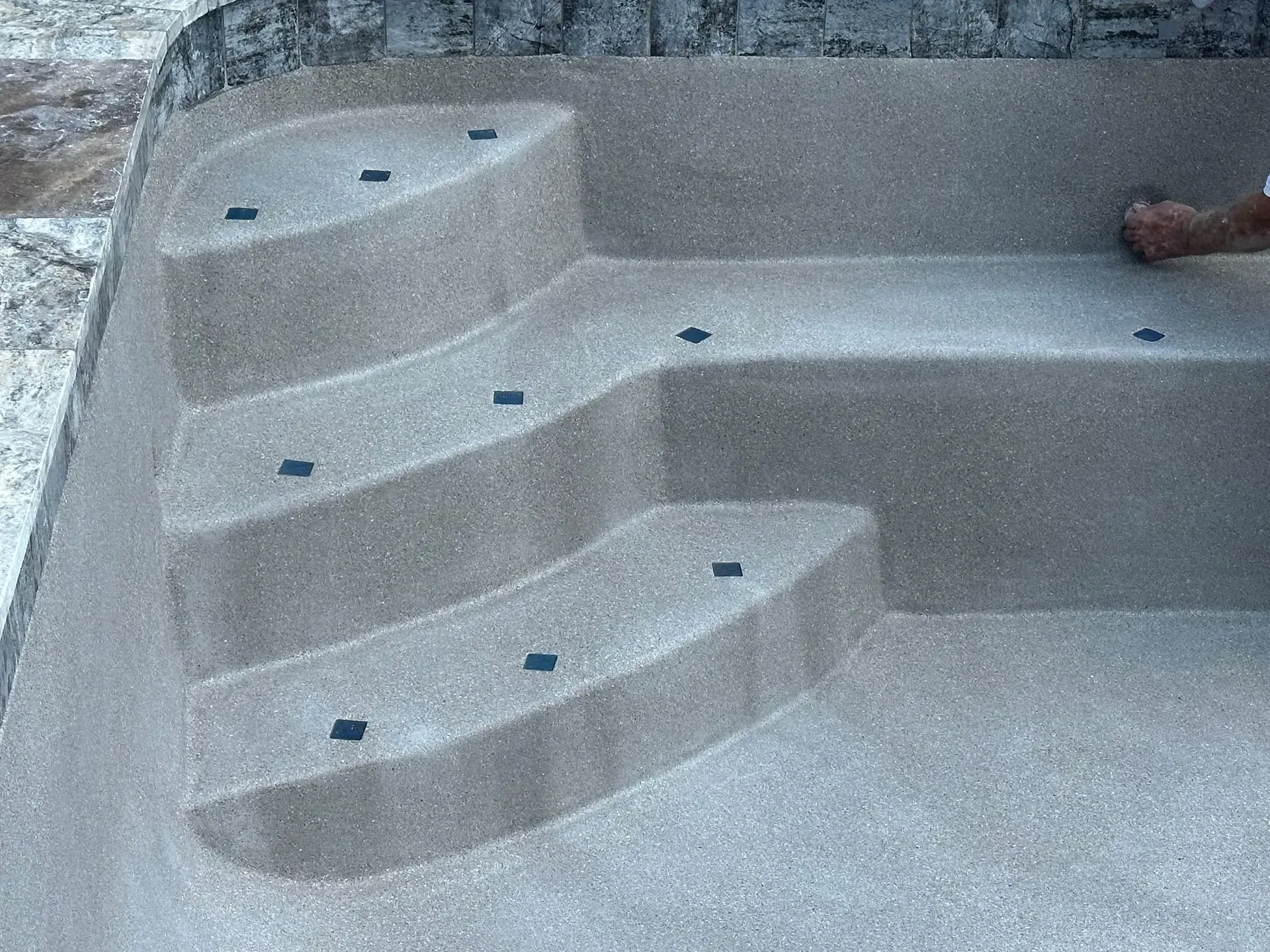 Pool steps with a textured, light-colored surface; person's hand on the edge.