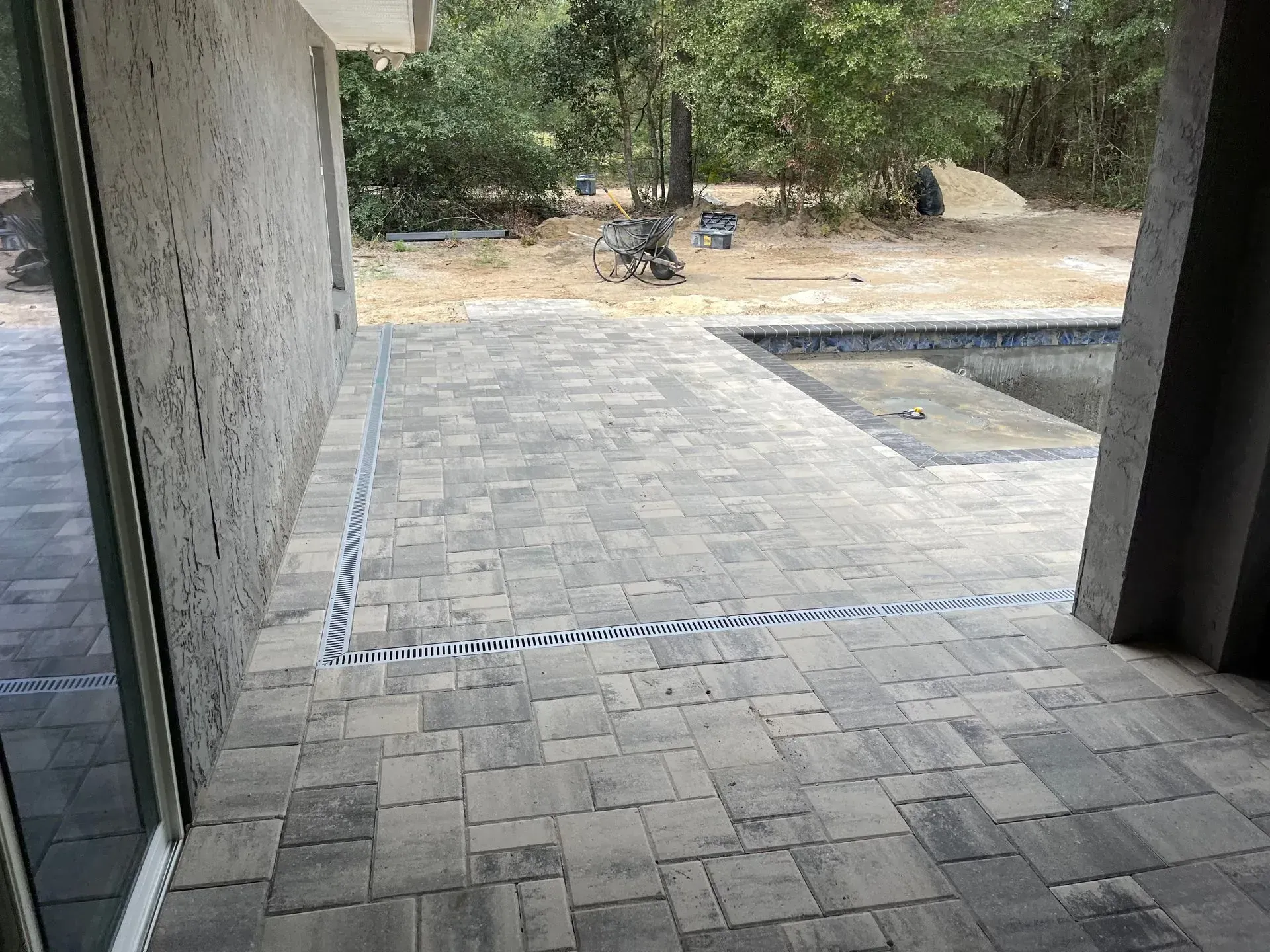 Stone patio with drainage channel, leading to a pool area and foliage.