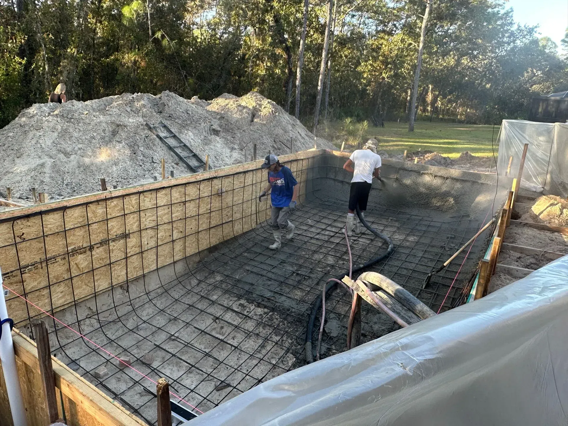 Construction workers spraying concrete into a pool's rectangular frame. Sunlight illuminates the outdoor setting with a pile of gravel nearby.