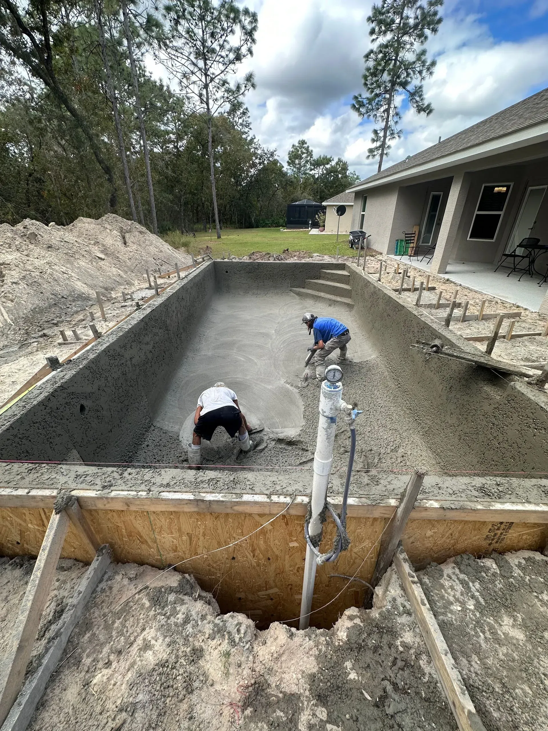 Two workers smoothing wet concrete in a rectangular pool under construction; gray and brown tones.