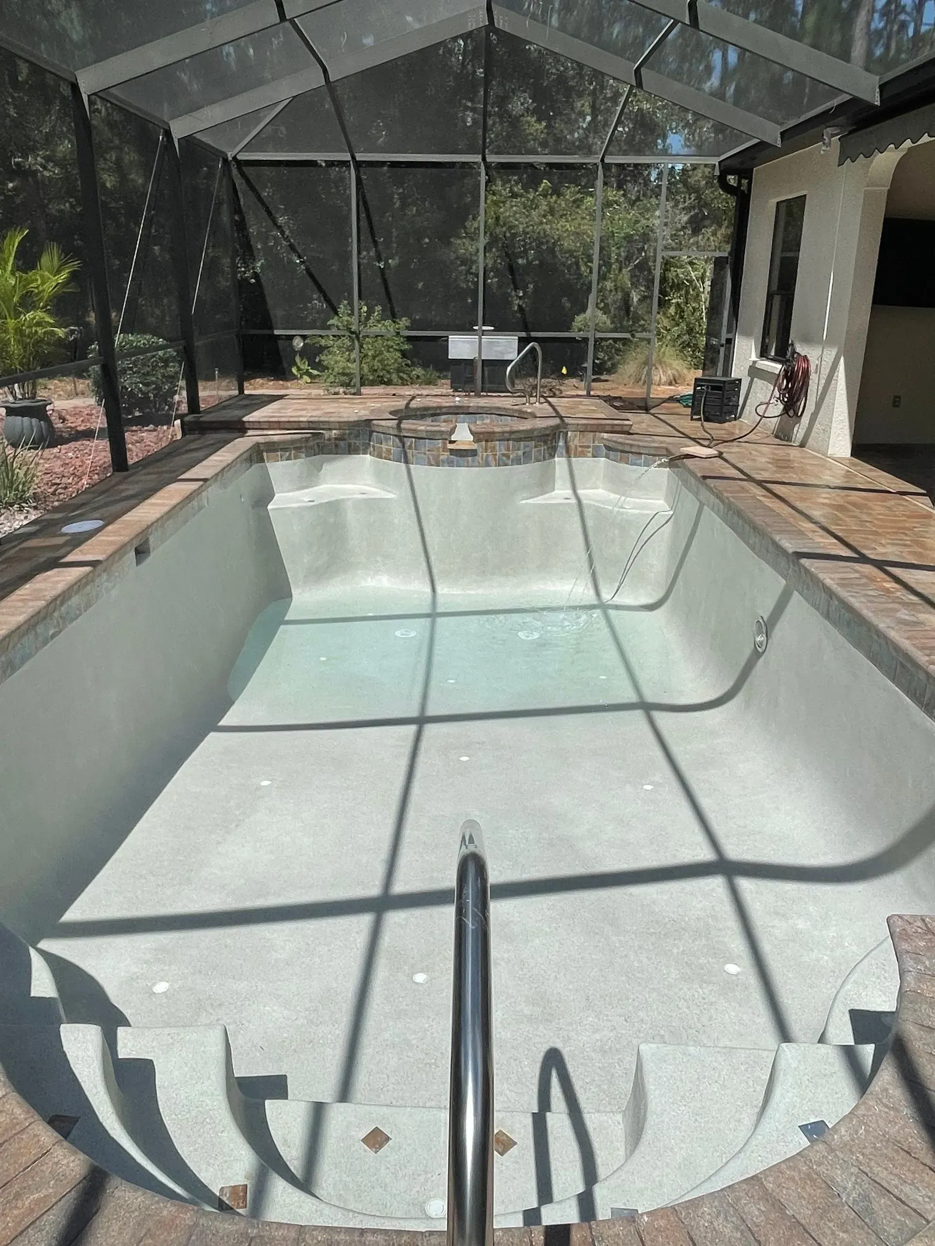 Empty swimming pool within a screened enclosure, steps on one end, brick patio surround.