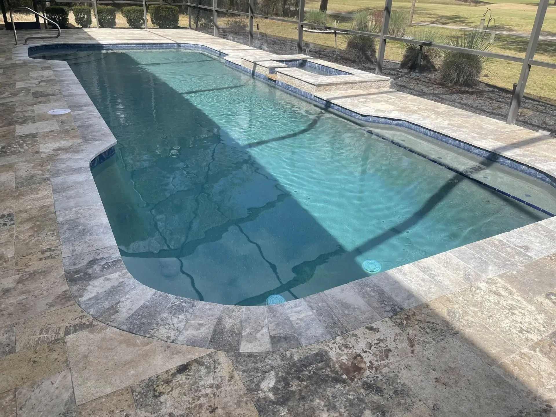 Pool with turquoise water, a hot tub, and stone patio surrounded by a fence.