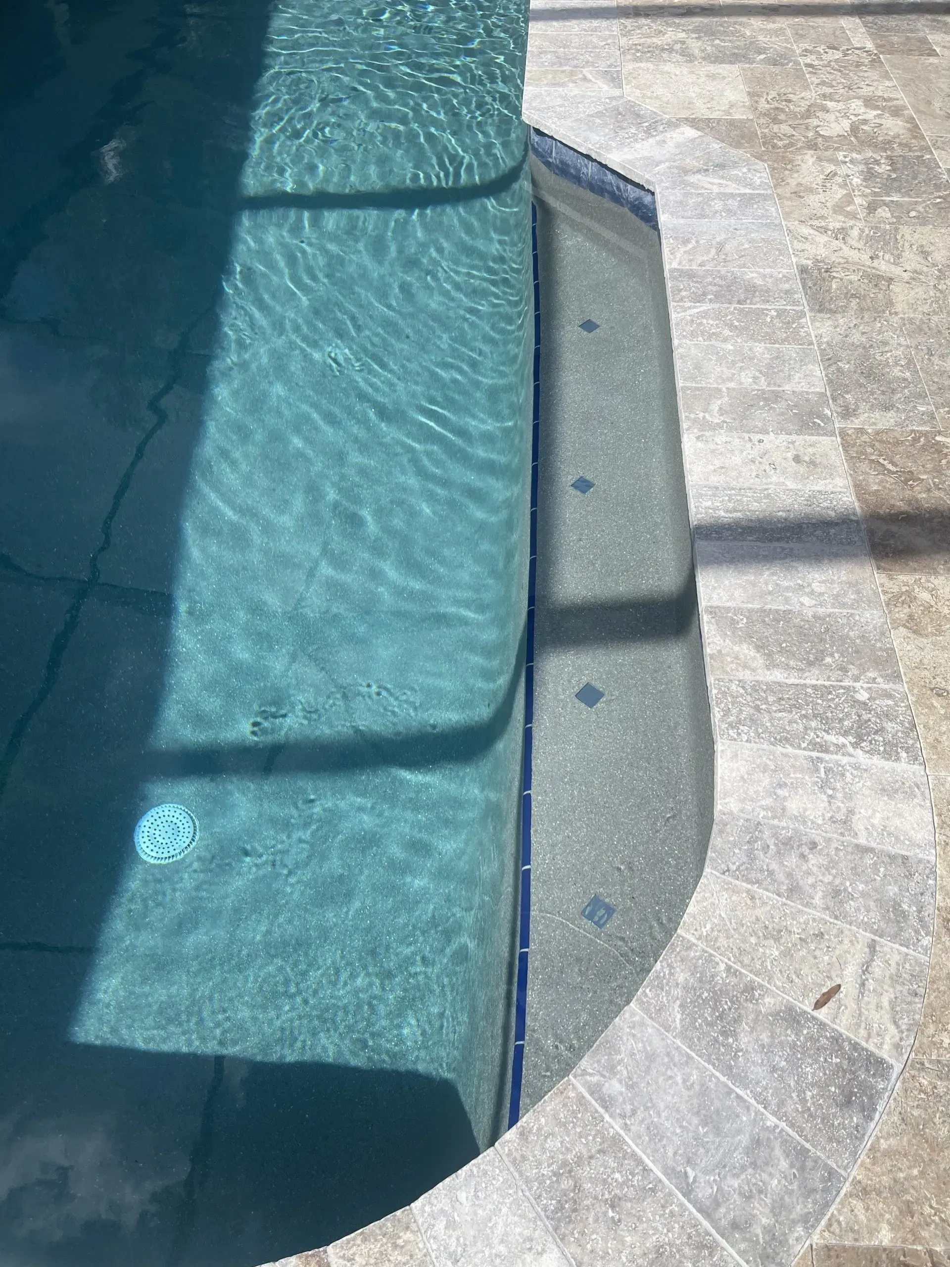 Pool steps with blue tiles and a stone deck. Sunlight casts shadows on the water's surface.