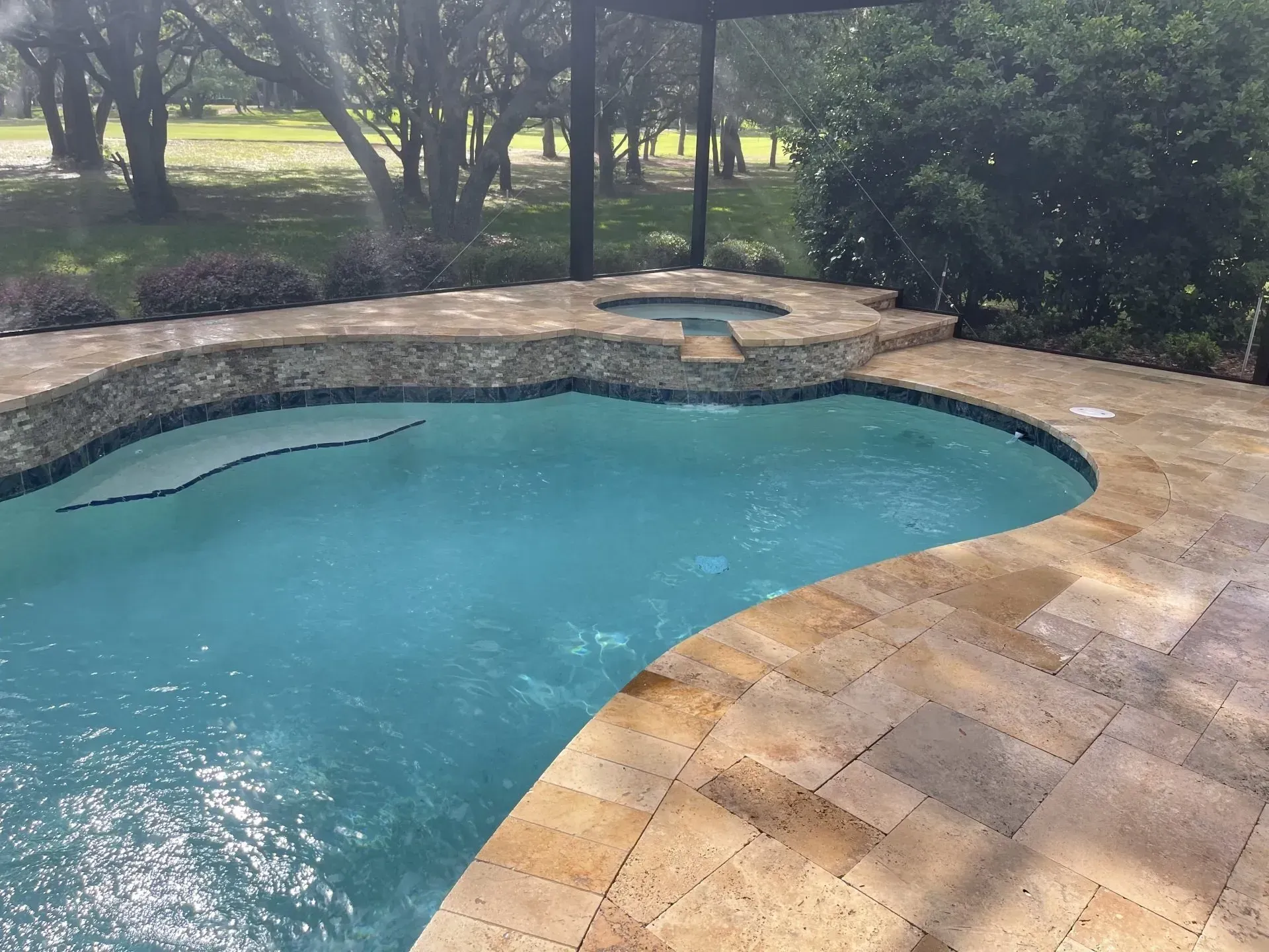 Swimming pool with attached spa, surrounded by stone patio, lush greenery in the background.
