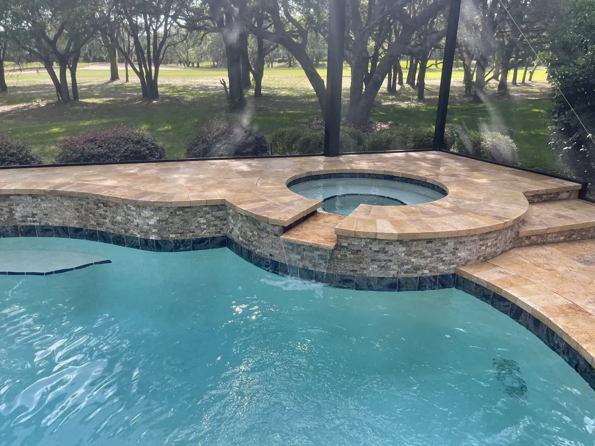 Swimming pool with attached spa and stone patio, trees in background.