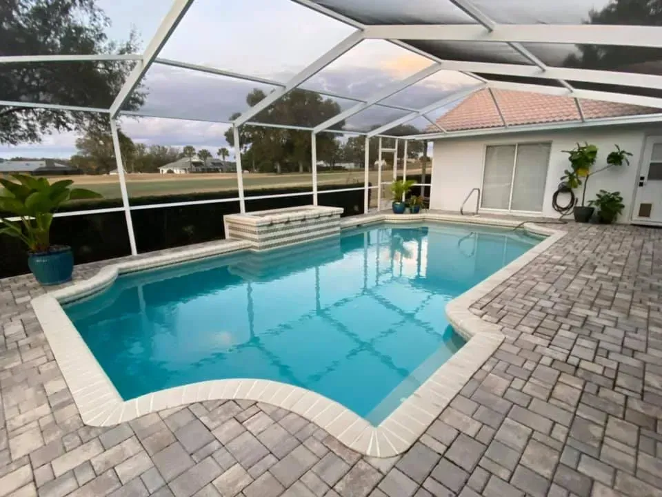 Swimming pool with screened enclosure and patio pavers, next to a white house.