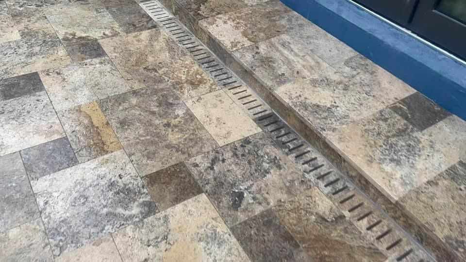 Tiled patio with a long, linear drain. The tiles are tan and brown. A dark blue edge is visible.