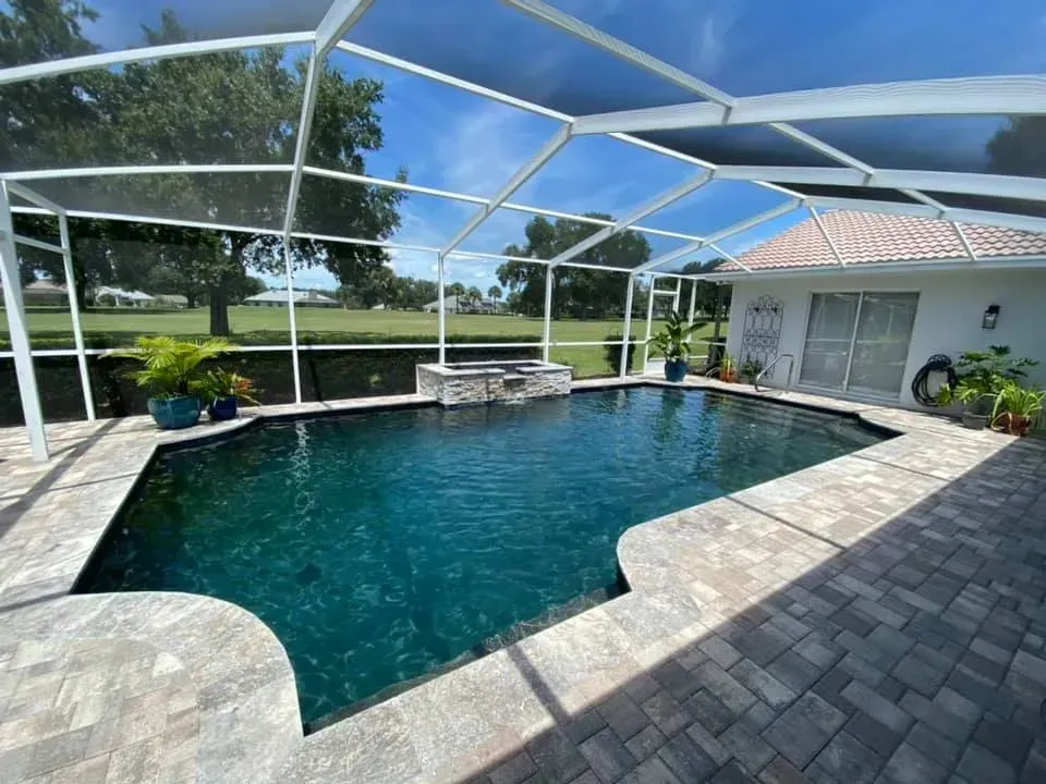 A screened-in swimming pool with dark blue water and stone patio, adjacent to a white house under a sunny sky.
