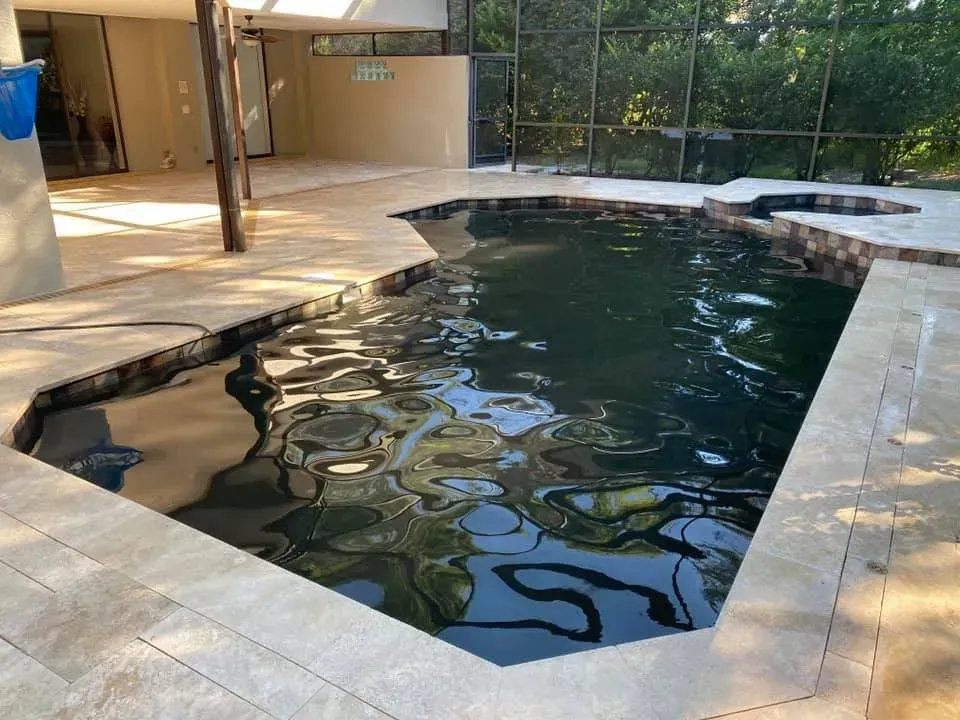 Pool with dark water, surrounded by light-colored stone patio. Open-air setting, adjacent to a building.