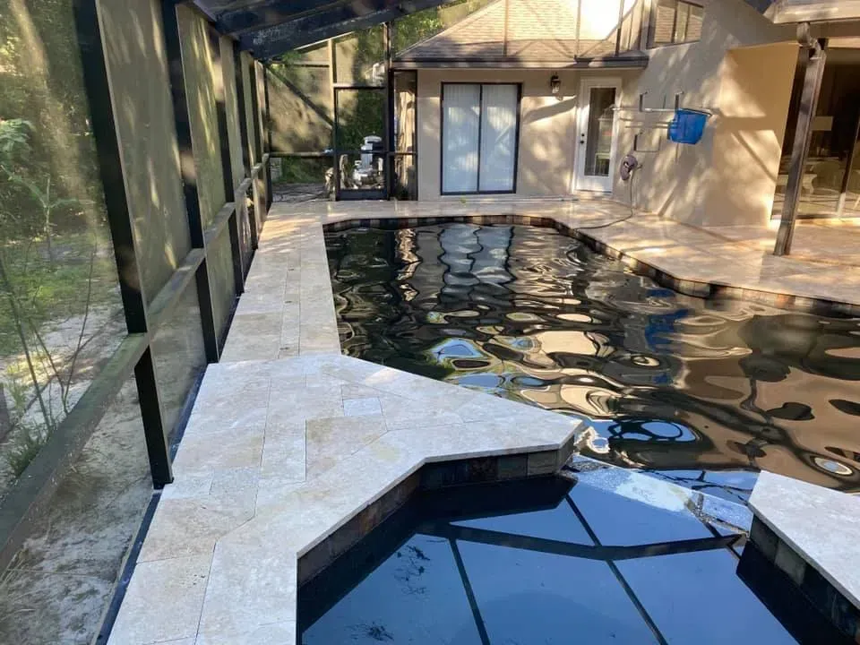 Pool and spa with beige tile surround, enclosed by a screen, next to a light-colored house.