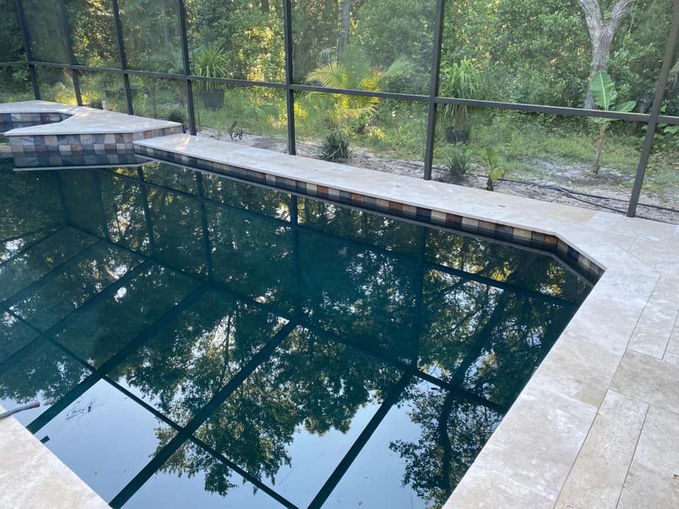Pool with dark water, surrounded by tan tile and a black screen enclosure, reflecting trees.