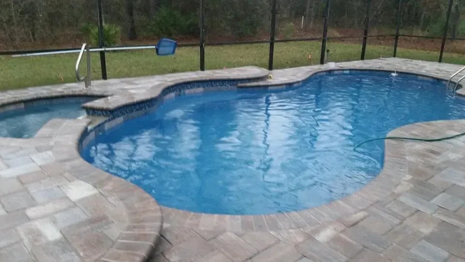 Blue swimming pool and hot tub surrounded by stone pavers, screened enclosure.