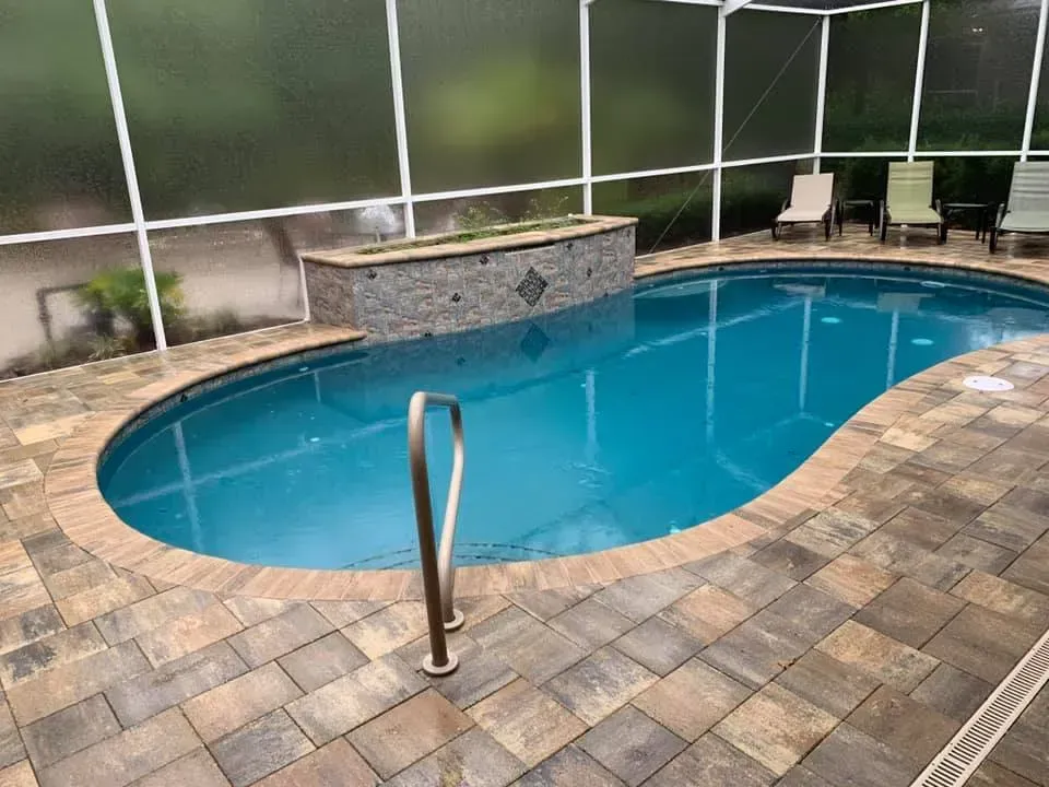 A swimming pool with brick patio, a small waterfall feature, and chairs inside a screened enclosure.