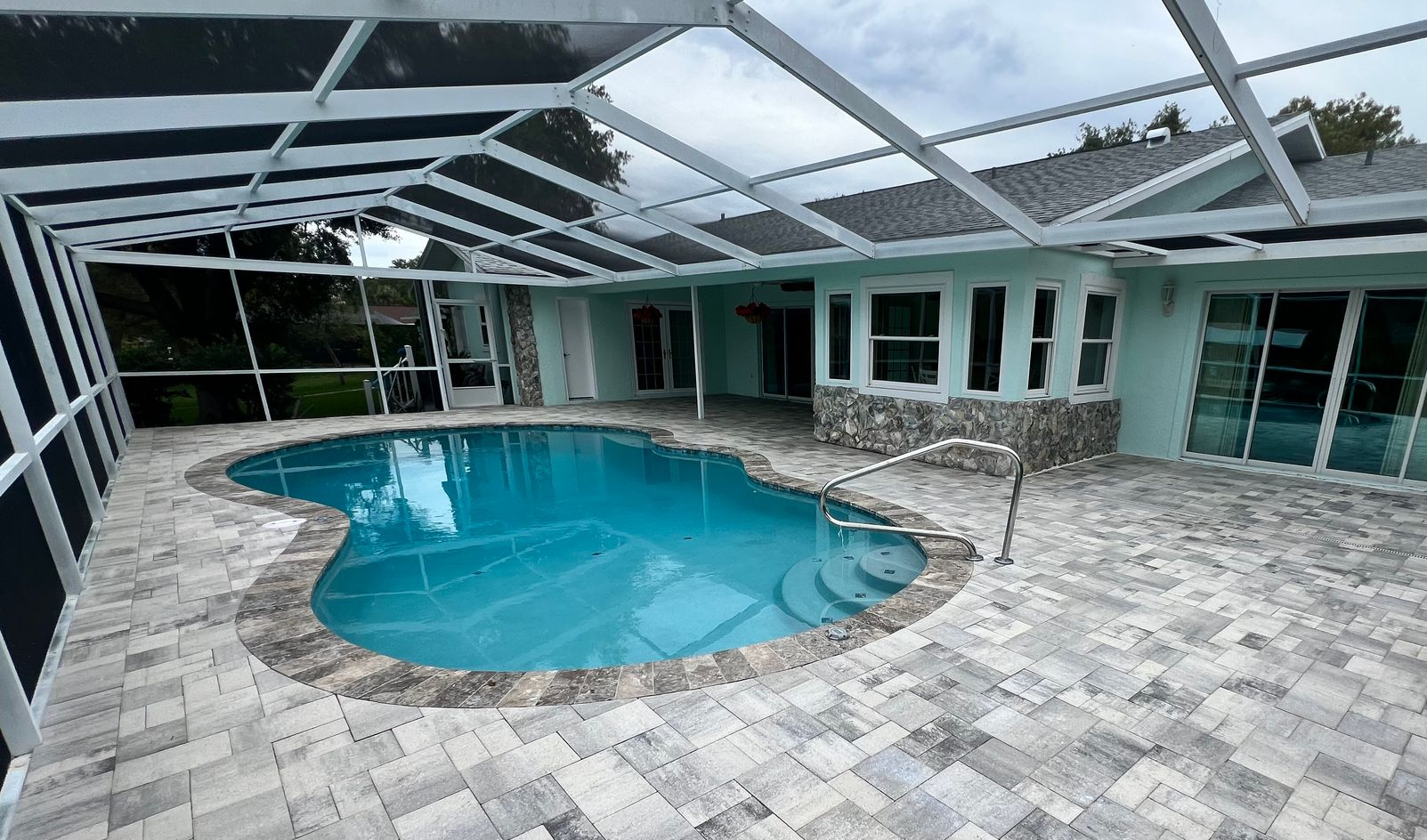 Screened-in patio with a turquoise pool and light stone pavers under a glass roof