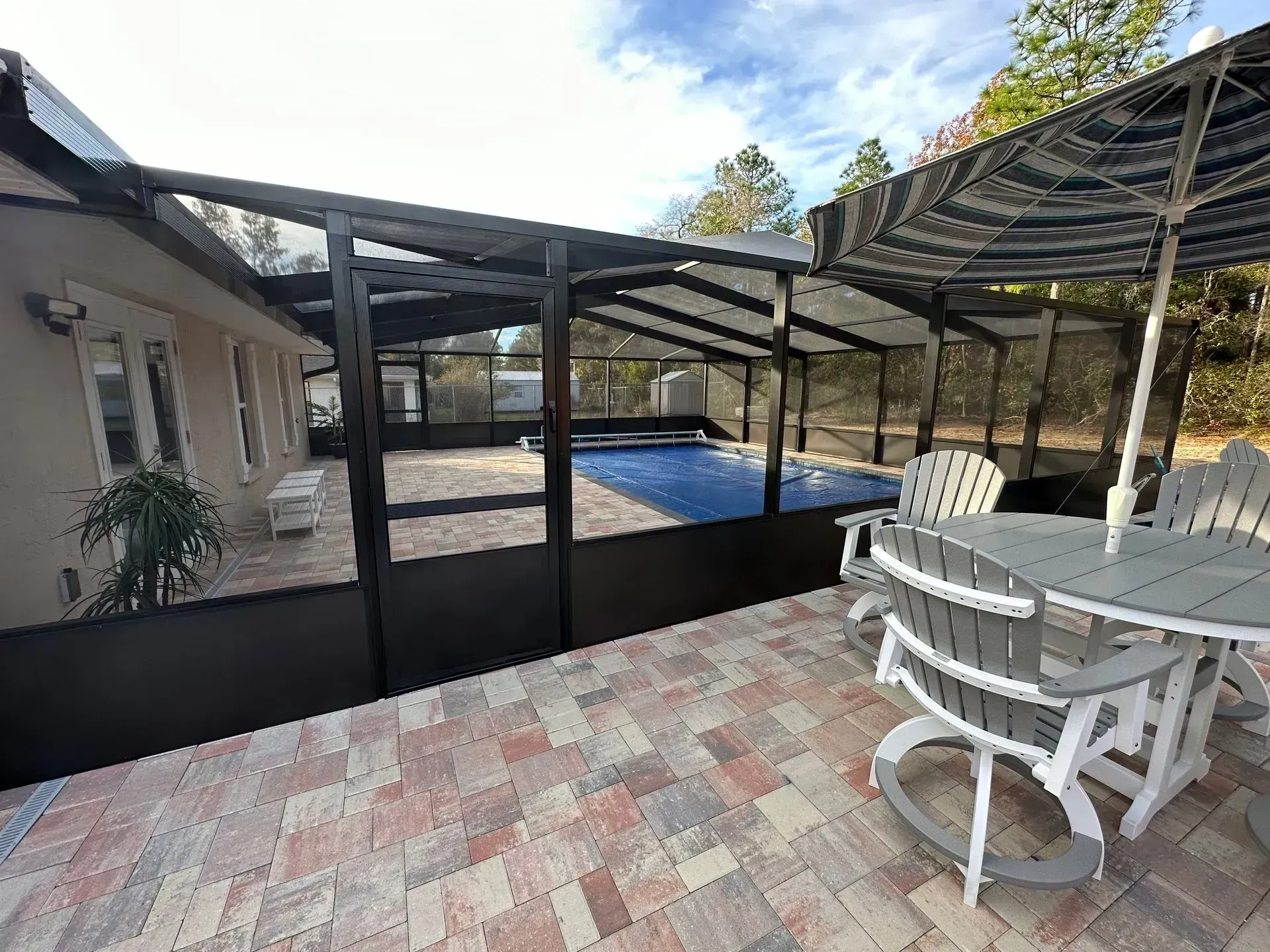 A paved pool deck enclosed by a dark screen, featuring patio furniture under an umbrella and a covered swimming pool.