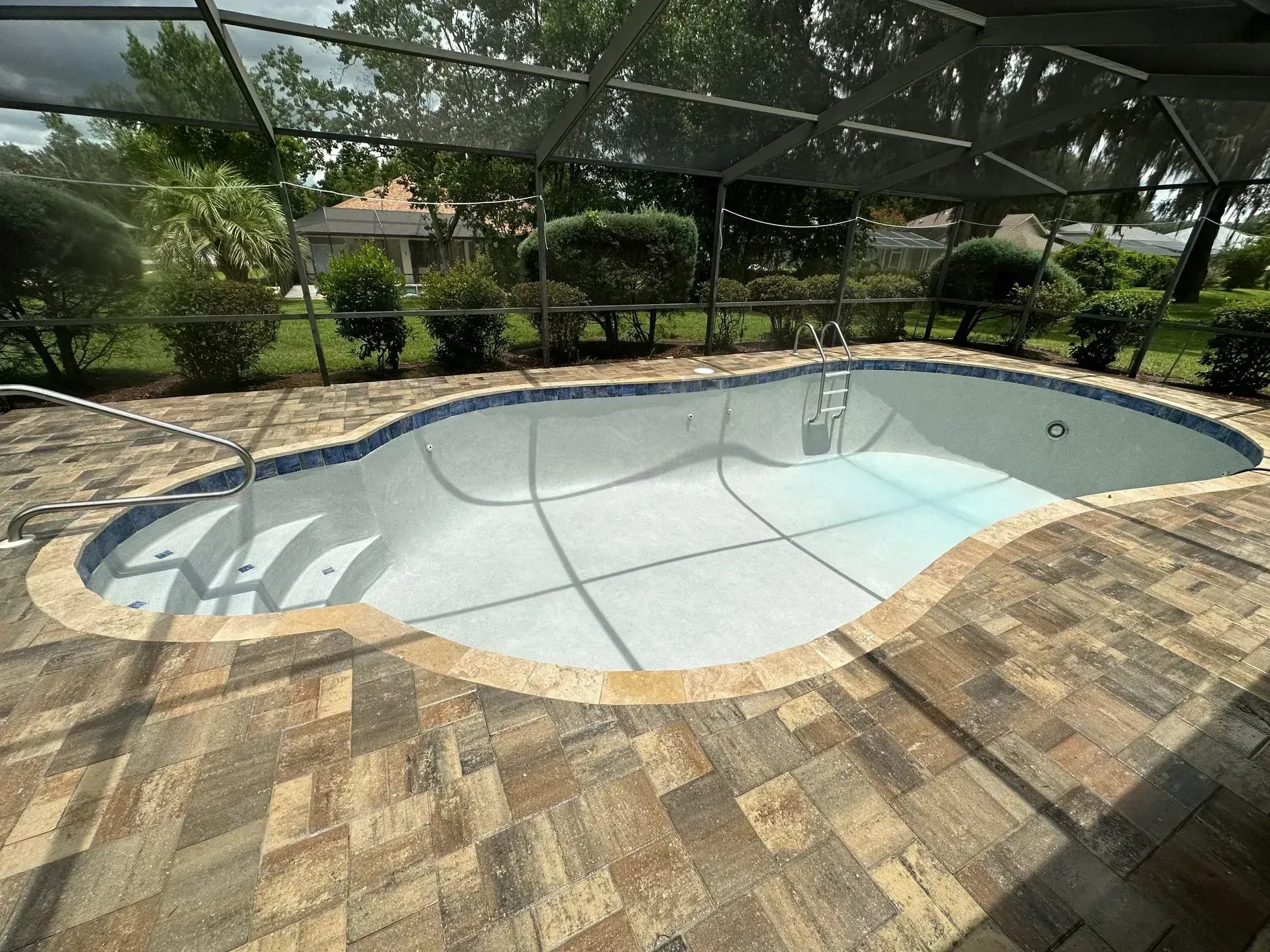 An empty, light-colored, kidney-shaped swimming pool with tiled edges, surrounded by brown stone pavers under a screen.