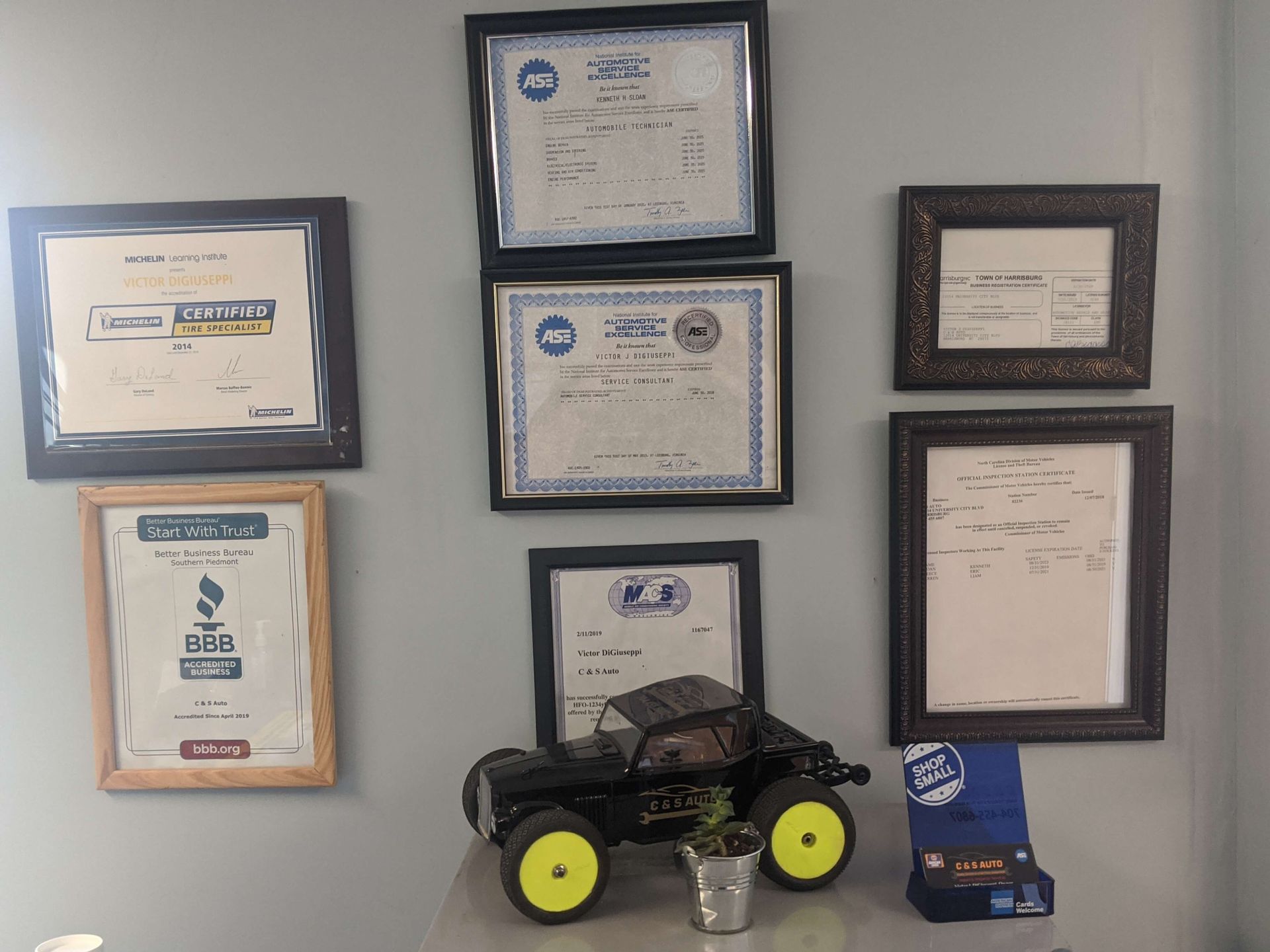 Wall of framed certificates above a remote-control car display with a small blue box on a shelf.