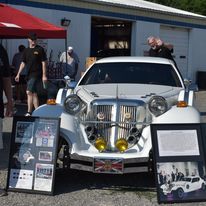 White custom car with chrome grille and yellow lights on display outdoors, with people and exhibit boards nearby