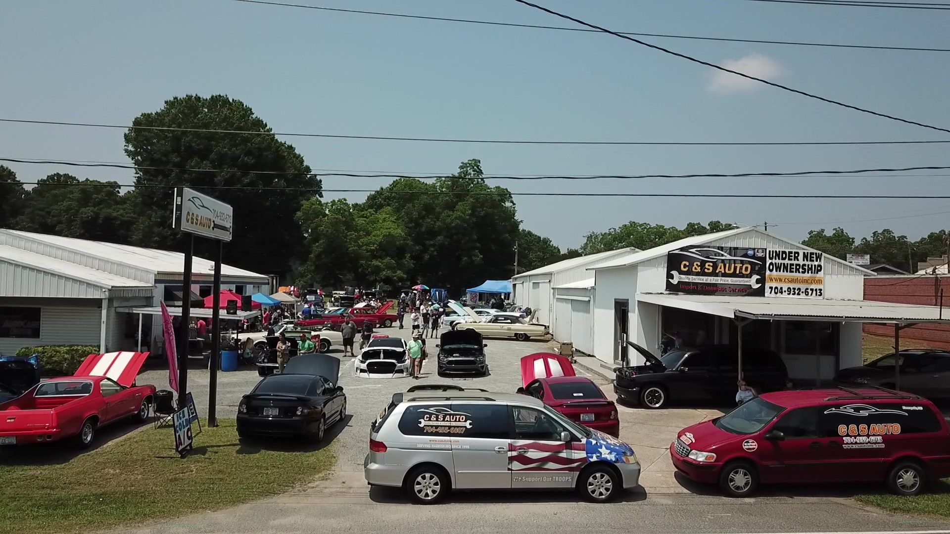 Outdoor car show with several parked cars and people milling around in front of white tents