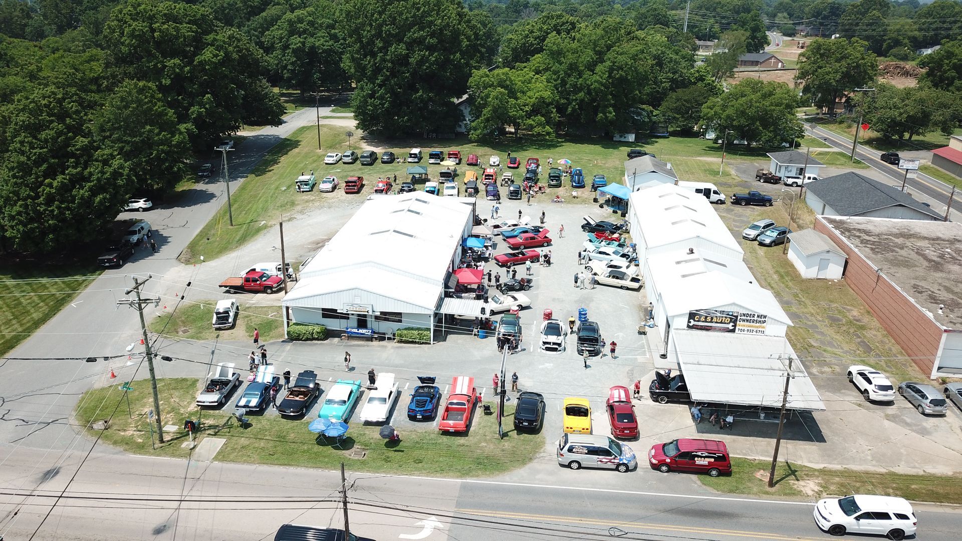 Aerial view of outdoor event tents, parked cars, and people gathered in a grassy lot beside a road.