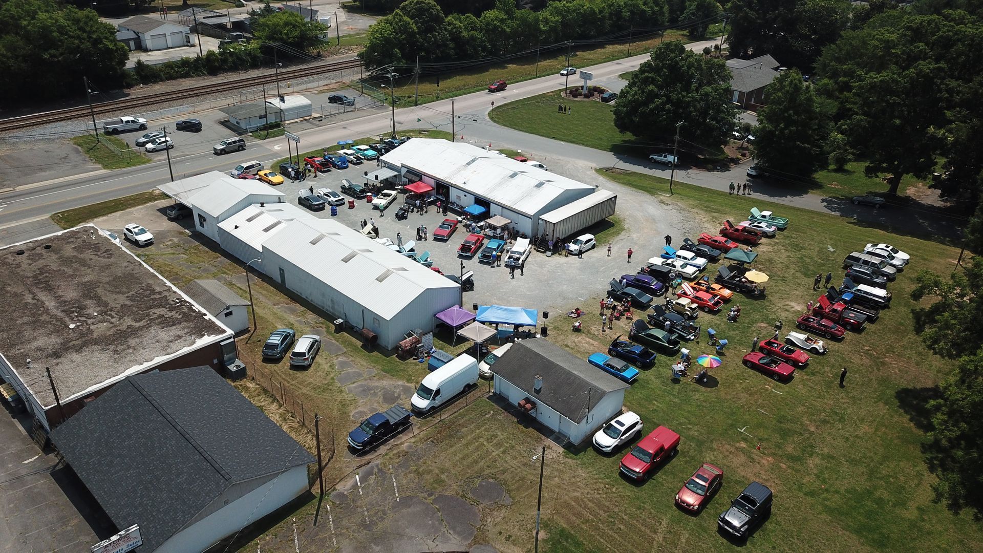 Aerial view of a community event with white tents, parked cars, and people gathered in a grassy lot.