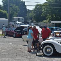 People standing beside a white vintage car at an outdoor car show, with other classic cars in the background