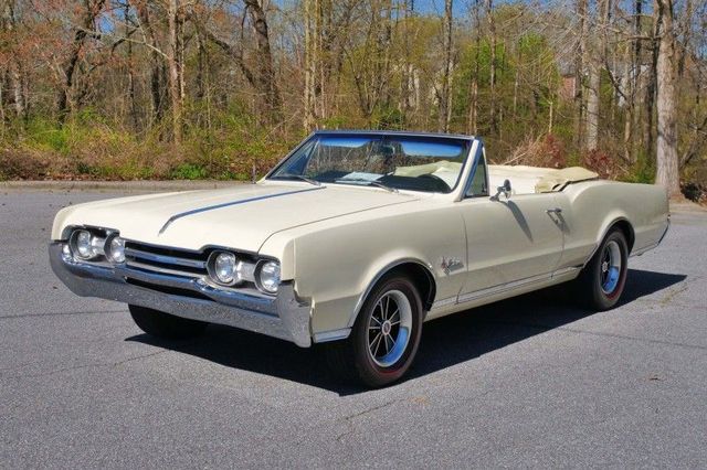 Cream-colored classic convertible parked on a roadside with trees in the background