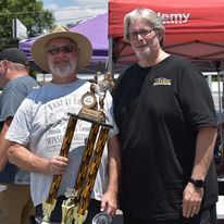 Two people posing outdoors, one holding a tall trophy, with red canopies in the background