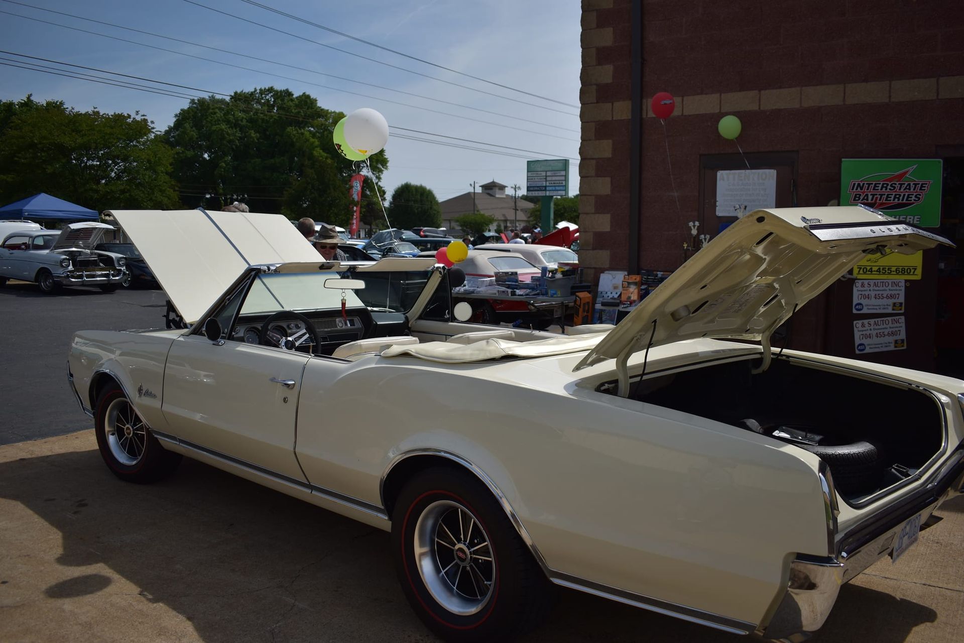 Classic white convertible at a car show with hood and trunk open, parked beside a brick building