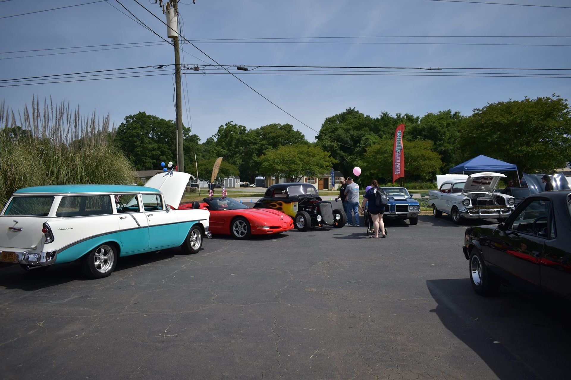 Classic cars lined up at an outdoor car show, with people gathered under tents on a sunny day