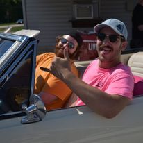 Two people in a cream convertible, smiling and waving, one in a pink shirt and cap, the other in orange sunglasses.