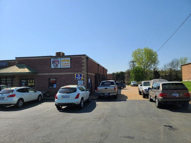 Small brick storefront with parked cars in a sunny lot, trees and blue sky overhead