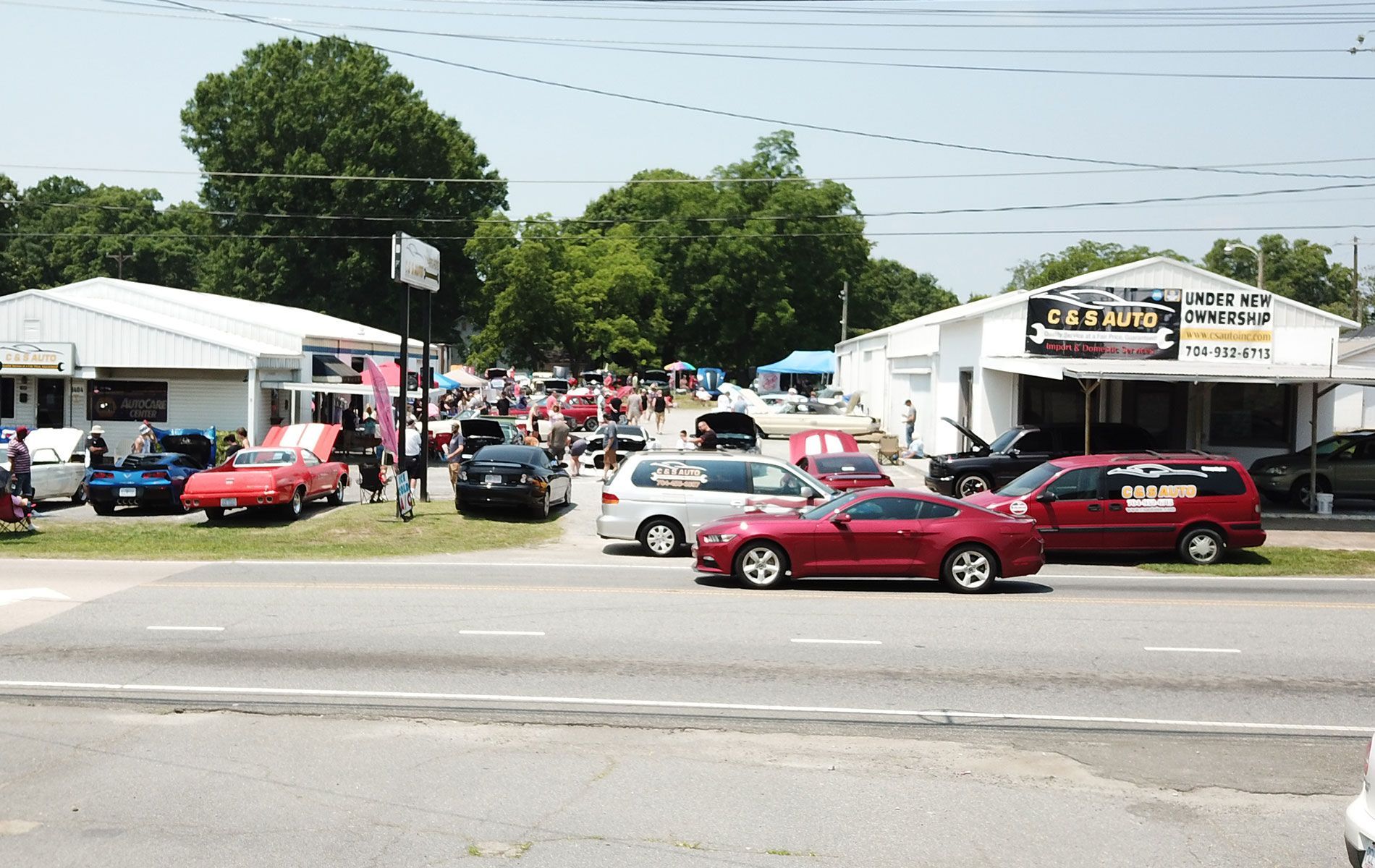 Car lot with rows of colorful cars and people gathered outside white tents on a sunny day