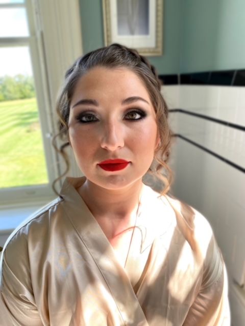 A woman wearing a robe and red lipstick is standing in front of a window.