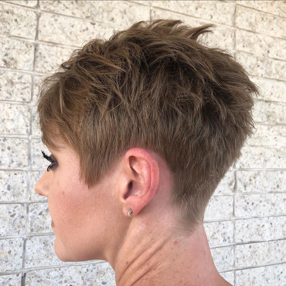 A woman with short hair and earrings is standing in front of a brick wall.