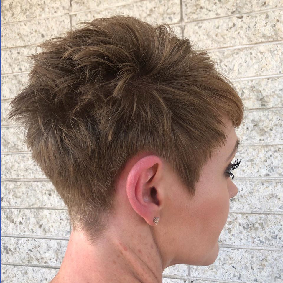 A woman with short hair and earrings is standing in front of a brick wall