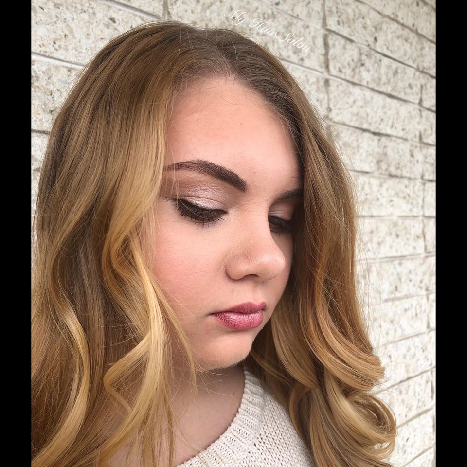 A woman with long blonde hair is wearing a white sweater and standing in front of a brick wall.