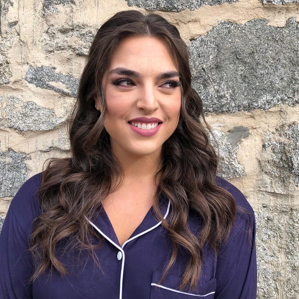 A woman in a purple shirt is smiling in front of a stone wall.