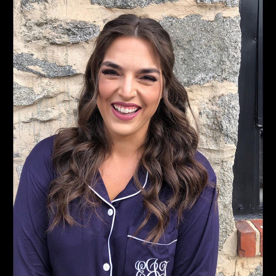 A woman in a purple shirt is smiling in front of a stone wall.