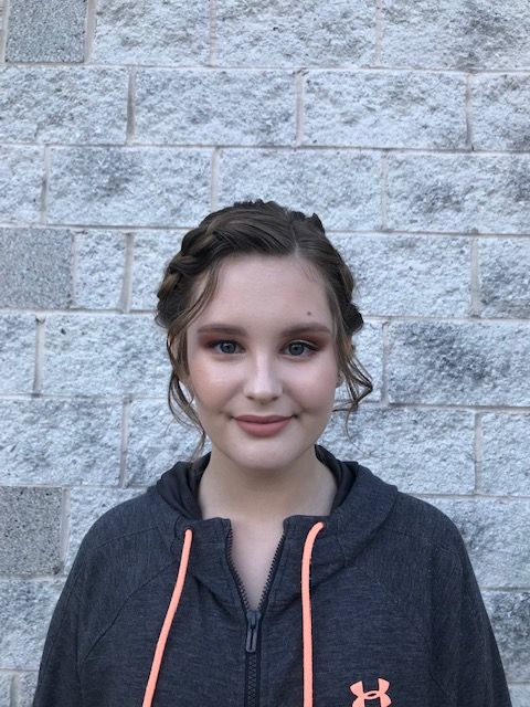 A girl wearing an under armour hoodie is smiling in front of a brick wall.