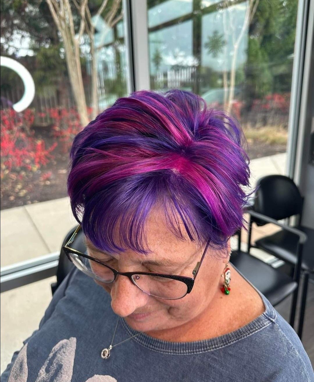 A woman with purple hair and glasses is sitting in front of a window.