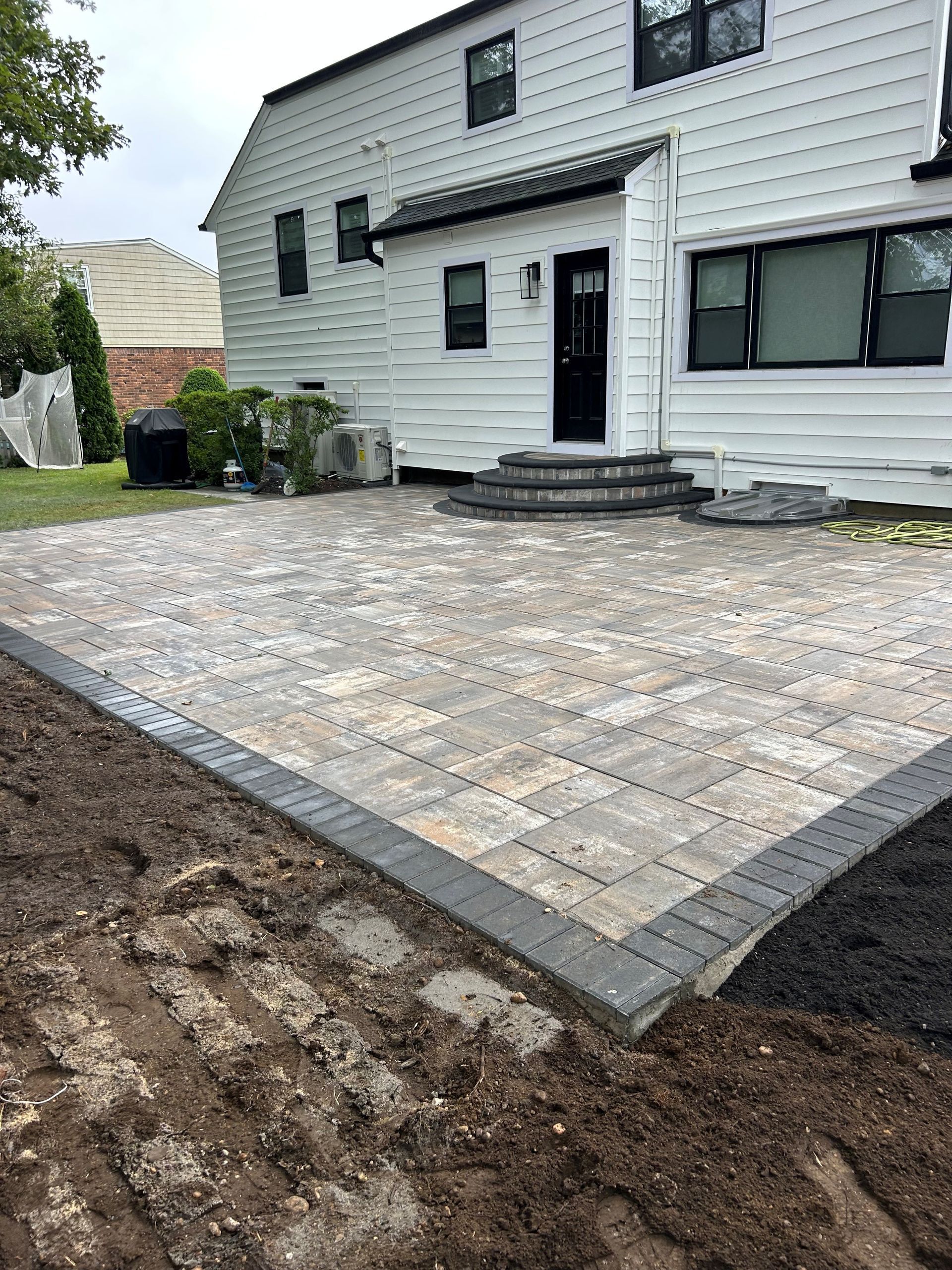 A newly constructed patio with pavers, gray border, and steps leading to a white house.