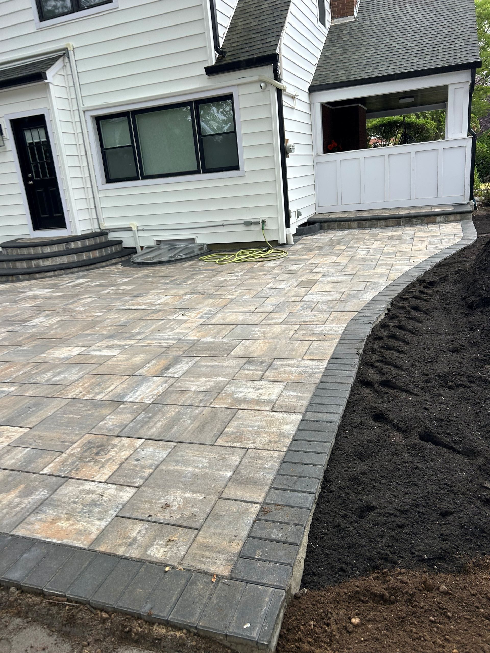 Stone paver walkway alongside a white house with black trim. Dark mulch border.