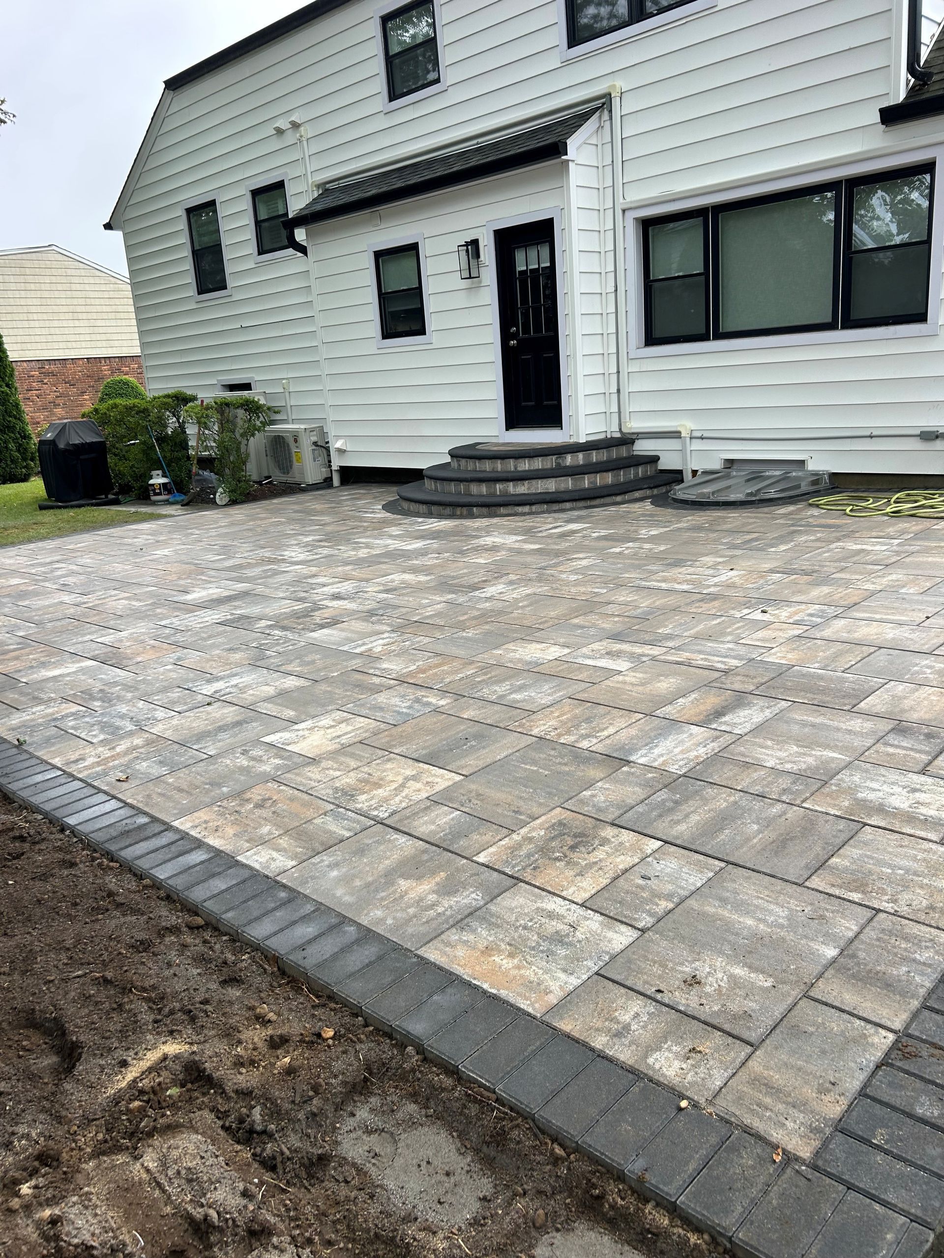 Backyard patio with gray and beige pavers, steps to a white house, and black trim.